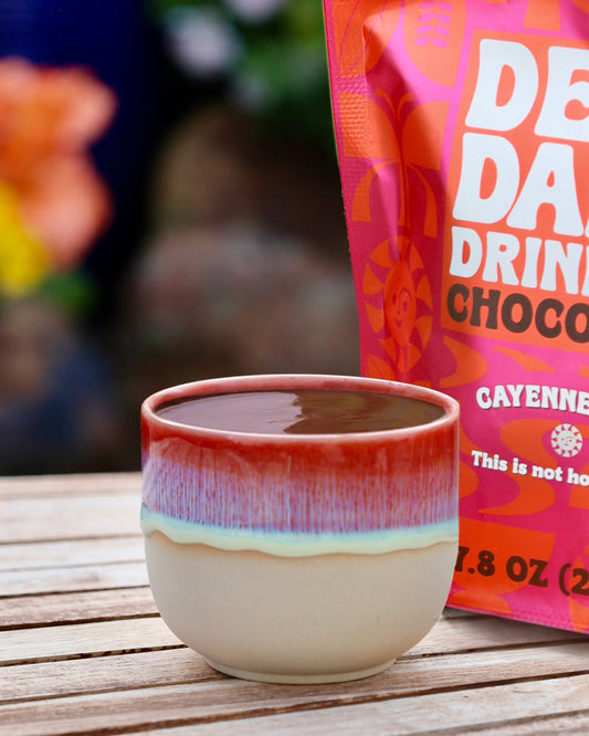 Ceramic cup and bag of Deep Dark Drinking Chocolate on a wooden surface with flowers in the background